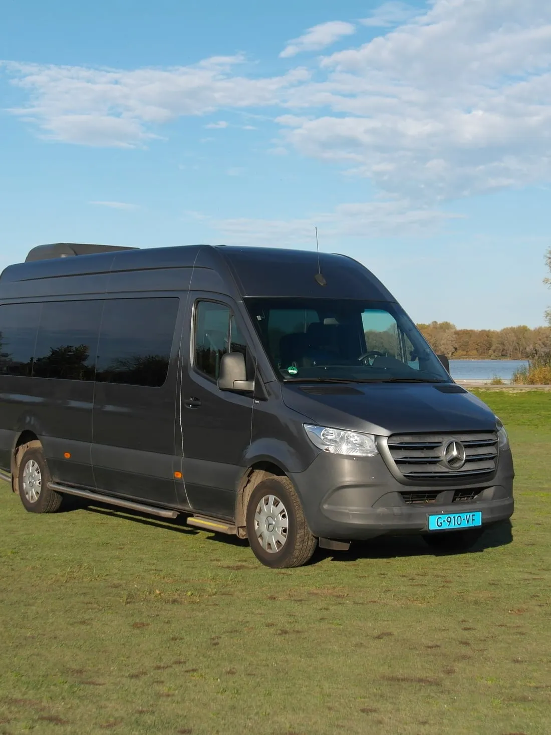 Grijze Mercedes Sprinter-minibus van Van Asselt, geparkeerd op een grasveld aan het water met bomen op de achtergrond